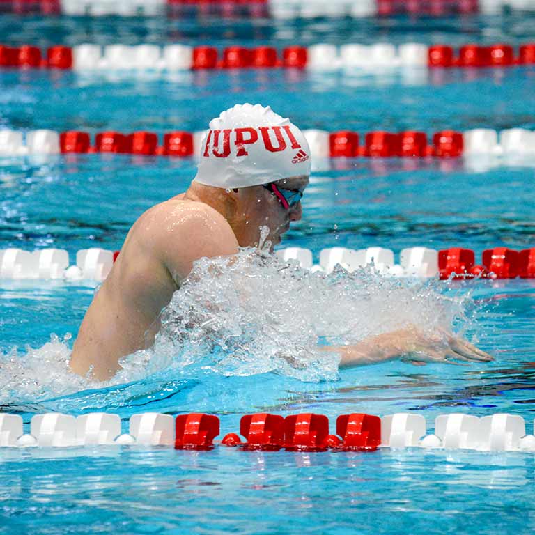 A male swimmer rises out of the water mid-stroke wearing an IUPUI swimcap.