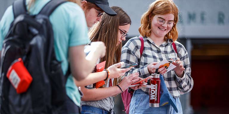 A group of students look at their phones, one student smiles while looking down holding a Weeks of Welcome water bottle.